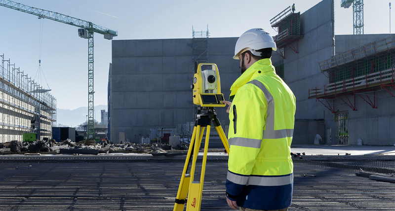 Työntekijä keltaisessa takissa ja suojakypärässä merkitsee linjoja ja pisteitä avoimella rakennustyömaalla.